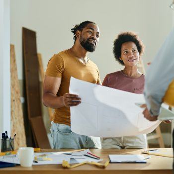 Happy African American couple analyzing blueprints while communicating with a building contractor in the apartment.